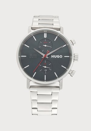 Montre-bracelet en métal argenté avec cadran noir, index horaires blancs, trotteuse rouge, deux sous-cadrans indiquant le jour et la date, et marquage HUGO.