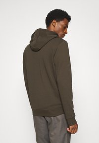 Felpa cappuccio marrone con polsini e orlo a coste, caratterizzata da un design semplice. Indossata con pantaloni a scacchi.