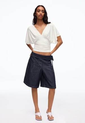 Donna che indossa un top bianco arricciato con scollo a V e maniche a pipistrello, pantaloncini larghi in denim scuro e sandali bianchi con tacco, in piedi con le mani dietro la schiena.