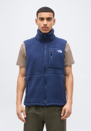 Ung mand iført en mørkeblå fleecevest med lynlåslomme på brystet og logo over en brun kortærmet skjorte.