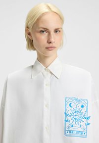 Een vrouw met kort blond haar die een witte blouse met knopen draagt, waarop een blauw tarotkaartontwerp staat met het label "De Minnaars" op de borst.