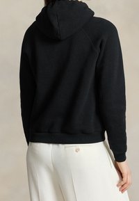 Černá mikina s kapucí, jemná textura, raglánové rukávy a žebrovaný lem. Nositelná přes bílé kalhoty, ukazující ležérní design.