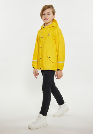 Giacca impermeabile gialla con cappuccio, bottoni a pressione, due tasche frontali, strisce riflettenti, indossata sopra jeans neri e sneakers bianche.