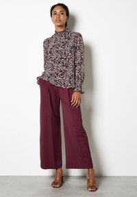 Donna con una blusa a maniche lunghe a fantasia, pantaloni larghi color bordeaux e sandali con tacco beige, in piedi contro un muro bianco.