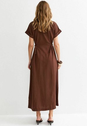 Mujer con cabello castaño claro ondulado, vestida con un vestido midi de manga corta marrón y sandalias negras con tacón, vista de espaldas sobre un fondo blanco.