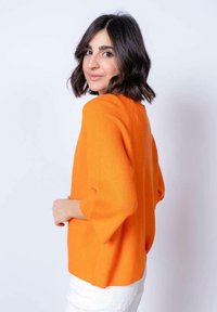 Maglione a coste arancione con maniche a campana a tre quarti, abbinato a pantaloni bianchi. Tessuto morbido e vestibilità comoda.