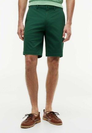 Mężczyzna noszący ciemnozielone szorty, jasnookie sweter i brązowe skórzane buty sznurowane, stojący na białym tle.