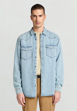 Hellblaues Jeanshemd mit verbreitetem Kragen, zwei Brusttaschen, Druckknöpfen und Kontrastnähten, getragen über einem beigen Hemd und sandfarbenen Hosen.