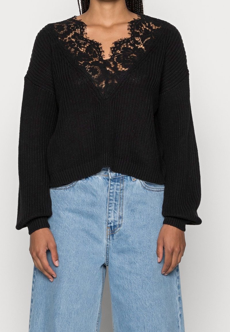 Pull en maille noire avec détail en dentelle au col en V, porté avec un jean bleu clair taille haute, visible des épaules jusqu'au milieu des cuisses.