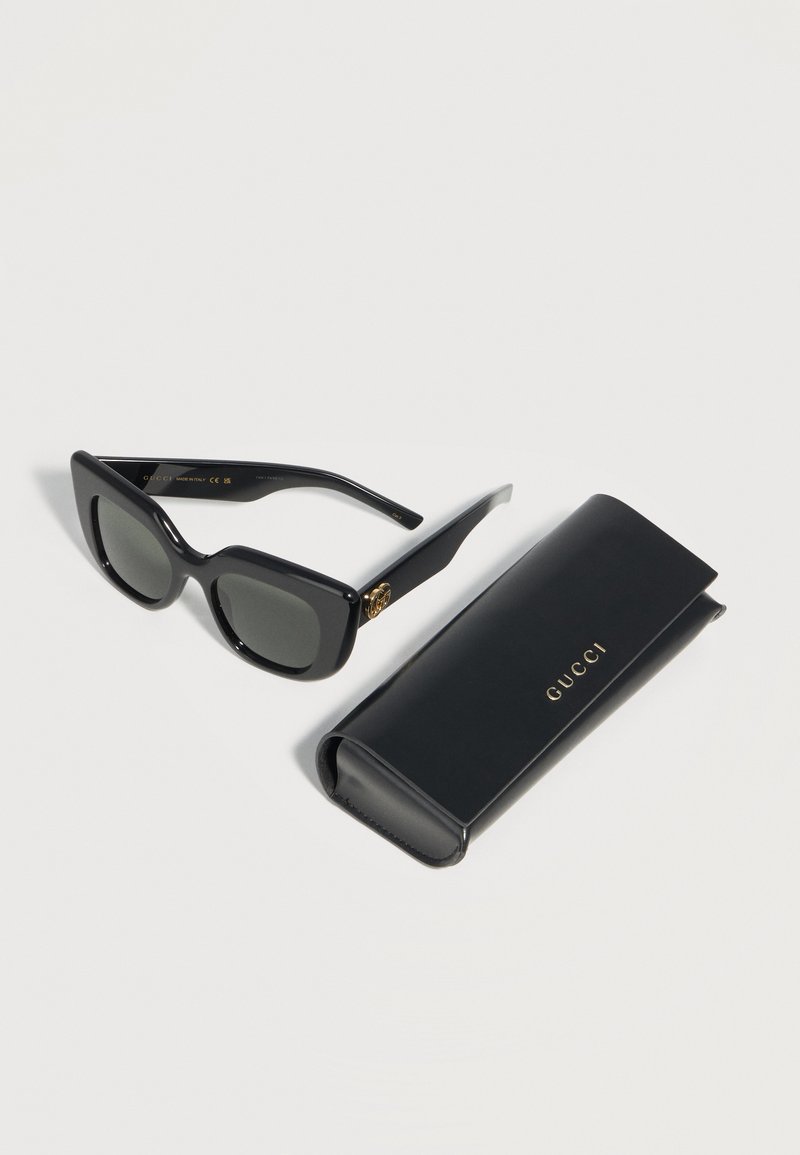 Occhiali da sole rettangolari neri con logo dorato sulle aste, accanto a una custodia nera Gucci abbinata con logo dorato su sfondo bianco.