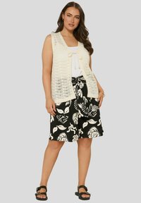 Stickad krämfärgad väst med öppet arbete, knuten fram, över en vit linne; i kombination med svarta shorts med vita bladmönster. Svarta platta sandaler.