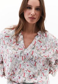Blusa floreale in tessuto leggero, con fiori rosa e grigi su sfondo bianco, scollatura a V e maniche larghi.