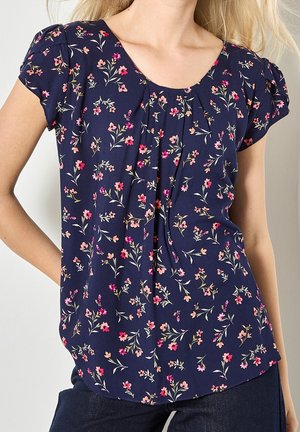 Femme portant un chemisier bleu marine à manches courtes avec un motif floral rose et orange clair, associé à un pantalon foncé.
