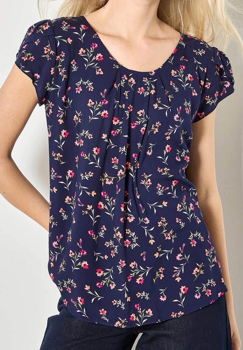 Femme portant un chemisier bleu marine à manches courtes avec un motif floral rose et orange clair, associé à un pantalon foncé.