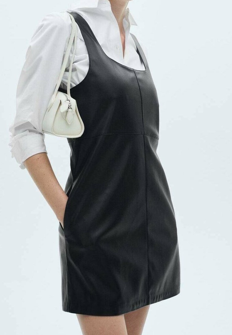 Femme portant une robe en cuir noire sans manches par-dessus une chemise blanche à manches longues, avec un petit sac à main blanc sur l'épaule.