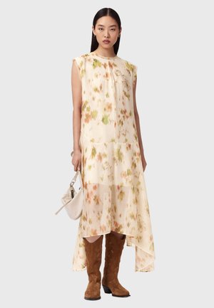 Frau trägt ärmelloses beiges Midikleid mit Blumenmuster und asymmetrischem Saum, braune Wildleder-Cowboystiefel und hält eine weiße Handtasche.