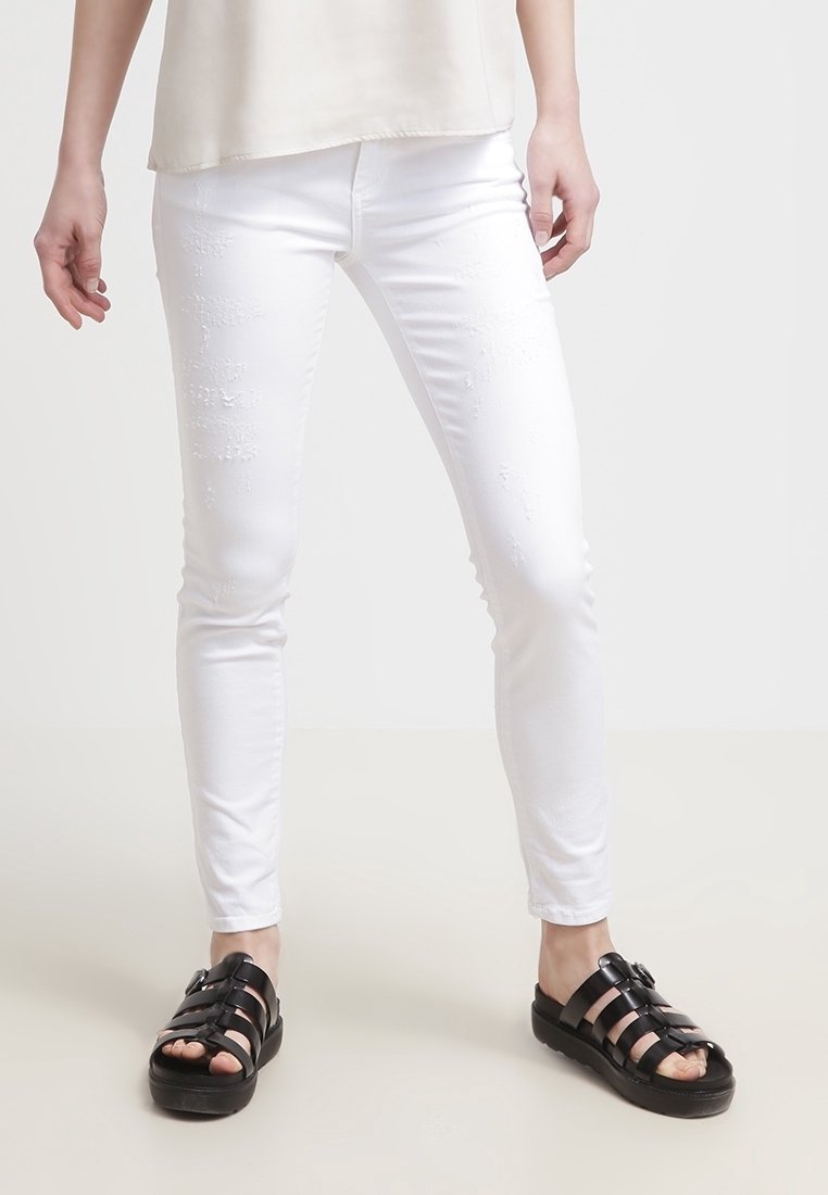 Personne portant un jean blanc slim déchiré et des sandales noires à boucle, debout sur un fond neutre.