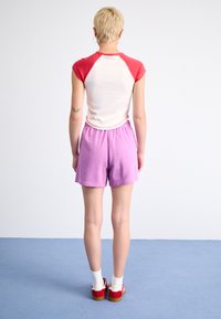 Kortermet skjorte med røde skuldre og hvit kropp, sammen med lilla shorts. Modellen har på seg røde sko med hvite sokker mot en blå bakgrunn.