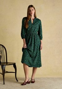 Roheline lilleline kleit, millel on kolmeveerandi varrukad, narmastatud talje ja põlvekõrgune hem. Seljas burgundia värvi plokknõelkingad. Lihtne, elegantne disain.