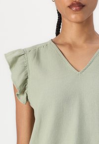 Top verde chiaro a V con maniche a sbuffo, realizzato in un morbido tessuto testurizzato. Presenta cuciture arricciate sulla cucitura della spalla.
