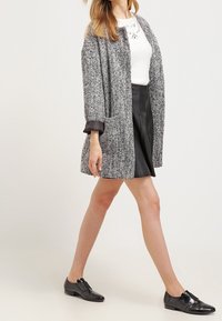 Manteau en tweed gris à coupe ample, manches longues et col. Porté sur un haut blanc et une jupe en cuir noir, assorti à des chaussures noires.