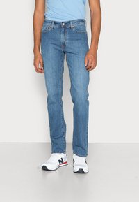Vaqueros de mezclilla azul claro con un corte recto, que cuentan con cinco bolsillos y costuras sutiles. Llevados con zapatillas blancas y una camisa azul.
