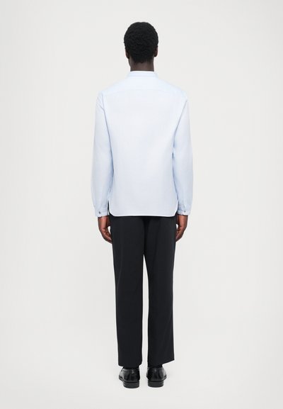 Jasnoniebieska koszula z długim rękawem o prostym kroju, z mankietami na guziki i z tylnym szwem, zestawiona z luźnymi czarnymi spodniami i czarnymi butami.