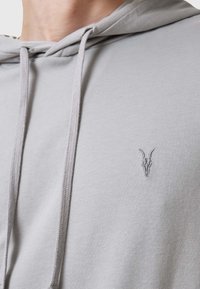 Sudadera gris claro con un bolsillo frontal en el centro, capucha ajustable con cordón y un pequeño logotipo bordado de un cráneo de cabra en el pecho.