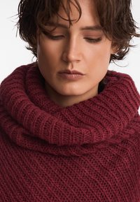 Bordowy sweter dziany z szerokim dekoltem typu cowl. Posiada teksturowany wzór, grubą przędzę i przytulny wygląd. Zbliżenie na detale materiału.