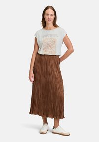 Jasnoniebieska koszulka z napisem "RADOSNY" i wzorem, zestawiona z brązową plisowaną spódnicą midi. Modelka nosi białe sneakersy.
