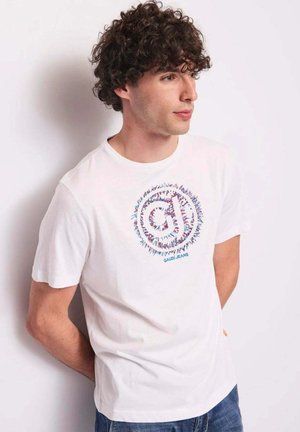 Giovane uomo con capelli ricci che indossa una maglietta bianca con un logo circolare colorato e jeans blu, guarda a destra contro una parete liscia.