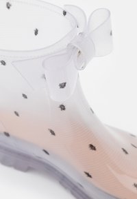 Transparent rain boot with black polka dots, curved shape, textured material, and a sheer bow accent at the ankle.