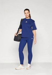 Γυναίκα που στέκεται σε μπλε αθλητική φόρμα Adidas με λευκές ρίγες, κρατώντας μια μαύρη τσάντα ώμου, φορώντας λευκά αθλητικά παπούτσια σε απλή φόντο.