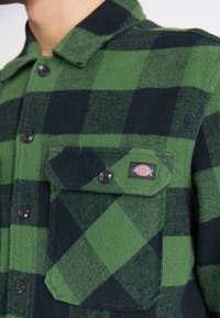 Grøn- og sortternet flanelskjorte med knaplukning fortil, sorte knapper, to brystlommer og et Dickies-logo patch. Blød tekstur.
