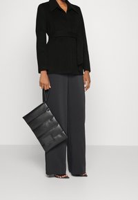 Chaqueta negra con cinturón y cuello, combinada con pantalones anchos negros. Se sostiene un bolso negro texturizado en una mano.