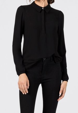 Kobieta ubrana w czarną bluzkę z długim rękawem, z małym kołnierzykiem i jednym widocznym guzikiem, w połączeniu z czarnymi spodniami.
