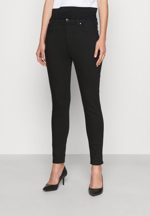 Frau trägt schwarze Skinny-Jeans und schwarze High-Heel-Schuhe, steht vor einem schlichten weißen Hintergrund.