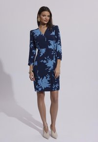 Granatowa sukienka z niebieskim kwiatowym wzorem, zawiązywana, z rękawami o długości 3/4, do kolan, w połączeniu z jasnymi, spiczastymi obcasami.