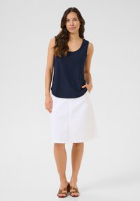 Haut sans manches bleu marine avec un motif texturé, jupe blanche à mi-mollet et sandales à enfiler marron, se tenant sur un fond uni.