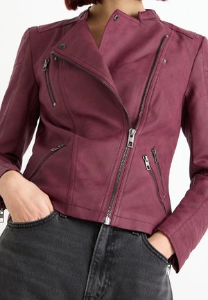 Personne portant une veste en cuir bordeaux asymétrique zippée avec plusieurs poches zippées et un jean noir taille haute.