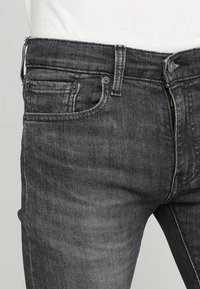Jeans de un gris oscuro con un ajuste Slim, que presentan costuras tonales, un diseño clásico de cinco bolsillos y herrajes metálicos en la cintura.