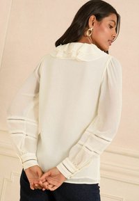 Blusa bianca con colletto a volant, tessuto trasparente, maniche lunghe a sbuffo e strisce ritagliate orizzontali sui polsini. Vista posteriore mostrata.