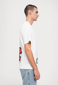 Camiseta de algodón blanca con un corte relajado, mangas cortas, que presenta un gran diseño gráfico en rojo y negro en la parte posterior.
