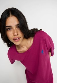 Fuchsiová tričko s krátkymi rukávmi vyrobené z bavlny, s okrúhlym výstrihom a malým modrým logom na hrudi. Hladká textúra, voľný strih.