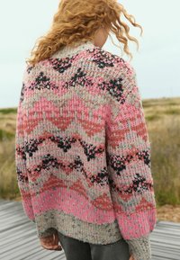 Pull en tricot avec un motif de vagues rose, gris et noir ; présente une coupe ample et un tissu texturé ; bords des poignets et du bas à côtes visibles.