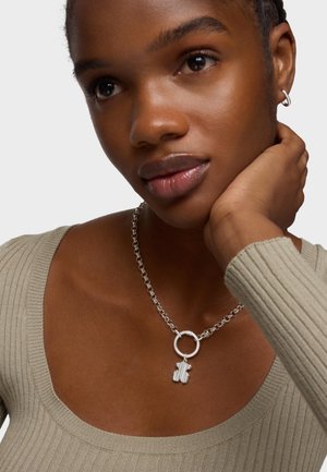 Collier en argent avec un pendentif en forme de main décoratif, relié par un charme rond, porté sur un haut beige à côtes avec un décolleté large.