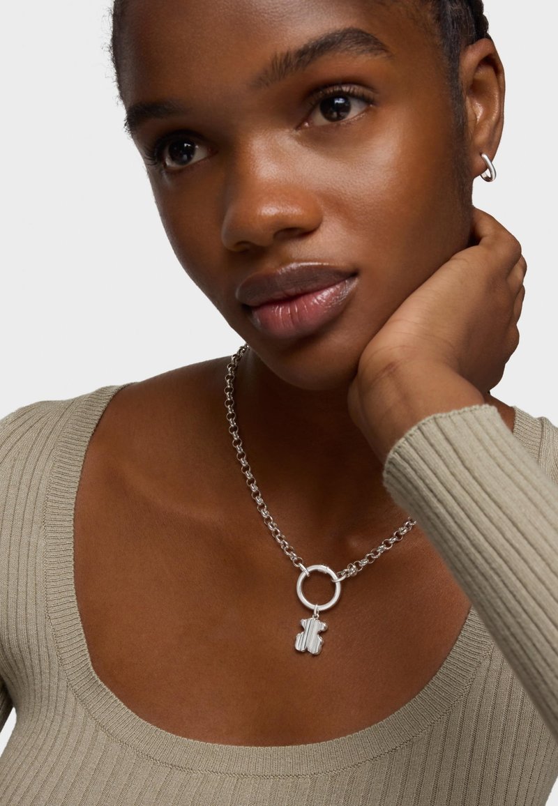 Collier en argent avec un pendentif en forme de main décoratif, relié par un charme rond, porté sur un haut beige à côtes avec un décolleté large.