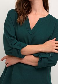 Teal skjorte med knaplukning, V-hals og trekvart ærmer, med rullede manchetter og et tekstureret stof.