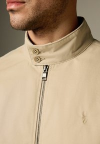 Gros plan d'une personne portant une veste beige avec un col montant, deux boutons, une fermeture éclair frontale et un petit logo cerf brodé sur la poitrine.
