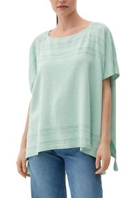 Mintgroene top met korte mouwen, met oogjesdetails en een losse pasvorm. Inclusief kleine franjes aan de zoom. Gedragen met blauwe jeans.